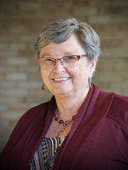 Head and shoulders image of Carol Harrison. She is standing in front of a brick background. She is wearing a red cardigan sweater. Her shirt underneath the cardigan has a white and black pattern with some pops of red. She is also wearing a red necklace and matching red earings which complement her cardigan. Her glasses have red frames which further complements her outfit.